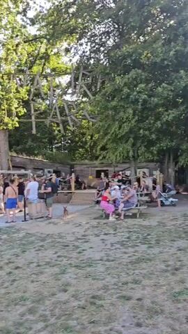 The video shows a park scene on a sunny day. In the foreground, a grassy area with some dirt patches is visible. In the background, there is a wooden structure with a sign that says "PARK" made of wooden letters. A group of people are gathered in front of this structure, some standing in a line, possibly waiting for something. A wooden building with a sloped roof is also present, and there are trees surrounding the area. Further into the park, there is a covered picnic area with tables and people sitting around them. The sky is blue and clear.