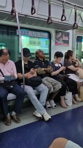 The video shows the interior of a public transport vehicle, likely a train or bus. Several passengers are seated on a bench. From left to right, an elderly Asian man with his left arm bandaged is looking down at a tablet or phone. Next to him, a young Asian person wearing a black bucket hat and glasses is looking at their phone. To their right, a man with long, light-colored hair, a beard dyed orange, and glasses is looking upwards with his arms crossed. He is wearing a black shirt with camouflage-patterned sleeves and camouflage pants. Next to him, a woman with short dark hair is looking at her phone. The last visible passenger is a woman with dark hair pulled back, also looking to her right. The seats are blue, and the floor is blue with white speckles. Handrails are visible above the passengers. A poster with Korean text and an image of a couple is on the wall behind the passengers. The overall impression is that of a typical commute.