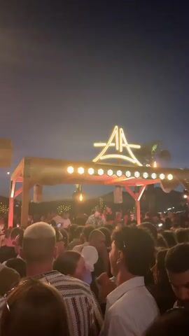 A nighttime outdoor party scene is captured. A crowd of people is gathered in front of a stage area. Above the stage, a neon sign with the letters "AA" in a stylized font is illuminated. The stage itself is wooden and has lights strung along its edge. A DJ, wearing a patterned shirt and glasses, is visible behind a console on the stage, appearing to be mixing music. Other individuals are seen on and around the stage. The sky is dark, indicating it is evening or night, with some distant lights and faint mountain shapes visible in the background. The overall atmosphere suggests a lively event with music and a gathering of people.