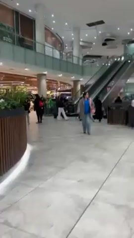 The video shows the interior of a large, brightly lit shopping mall, with a wide, marble-tiled floor and multiple levels visible. There are several people walking through the mall, some individually and others in groups. The crowd includes people wearing a variety of clothing. There is an escalator, and a portion of a supermarket with fresh produce visible in the background.  Various retail store signs are visible along the upper levels. The camera moves as the videographer walks.
