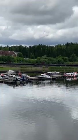A vertical video clip shows a calm body of water with a marina on the right side. Numerous boats are docked, with several boats of varying sizes visible. The water reflects the cloudy sky and the docked boats. In the background, a line of trees forms a dense forest, and beyond that, buildings and a road are visible. The sky is overcast with dramatic cloud formations. There are no people clearly visible in the clip. The overall scene depicts a serene marina environment on a cloudy day.