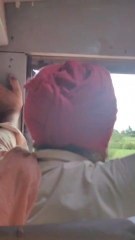 The video shows a close-up, rear view of a person wearing a red turban. The person is sitting in a vehicle, leaning out of a window. The background shows a landscape of green fields and trees under a cloudy sky. The person's right arm is visible, resting on the edge of the window. Another person's hand is visible on the left side of the frame, also near the window. The interior of the vehicle is dimly lit. The video is shot from a low angle, looking up at the person.