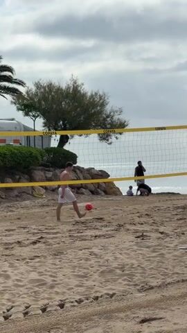 A video shows a beach scene with a volleyball net in the foreground. A shirtless man in white shorts stands near a red ball on the sand. The camera pans right, revealing two wooden posts supporting the volleyball net. The ocean is visible in the background under a cloudy sky. Another man in a black and red shirt holding a ball stands near the net, looking towards the right. Two other people are also visible in the distance, closer to the ocean.