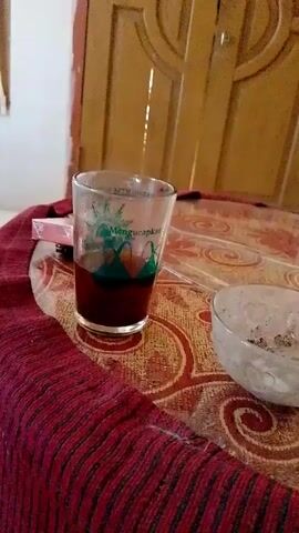 The video shows a glass of dark liquid, possibly coffee or tea, sitting on a patterned tablecloth. Next to the glass is a small ashtray containing cigarette butts. In the background, a wooden door with decorative carvings is visible. There are no people or obvious actions taking place in the scene. The lighting appears to be artificial.