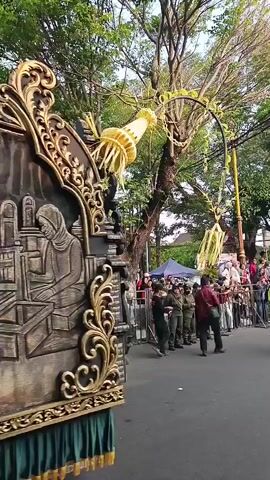 The video shows a close-up of a large, ornate float with intricate relief carvings. The float appears to be part of a parade or festival. The carvings depict scenes of people engaged in various activities, possibly historical or cultural in nature. One carving shows a woman in a headscarf sitting at a desk with books. Another shows a man in a suit sitting at a desk with a typewriter. The float is decorated with gold trim and green fabric at the bottom. In the background, trees are visible, and a crowd of people, some in uniform, are gathered. Yellow decorations are hanging from a tree branch above the float. The overall impression is of a vibrant cultural event.