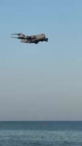Here's a brief description of the video:

A gray U.S. Air Force C-17 Globemaster III transport plane is seen flying relatively low over the ocean on a clear, blue sky day. The plane appears to be in landing configuration, with its landing gear deployed. A small red buoy is visible floating on the surface of the water below.