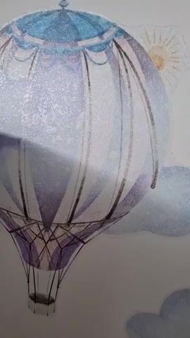 A video shows a close-up of a wall decal featuring a hot air balloon. The balloon is depicted in watercolor style, with shades of blue, purple, and white. It has a basket attached and is surrounded by clouds, stars, and a sun. The decal appears to be applied to a plain white wall.
