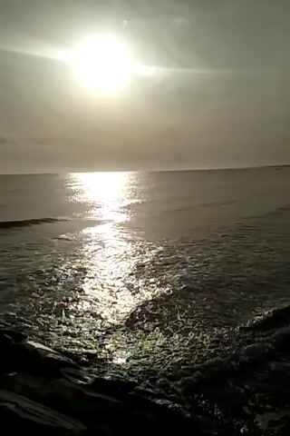 The video begins with a shot of the sun shining brightly over the ocean, casting a shimmering reflection on the water. Waves crash against a rocky shore in the foreground. The camera then pans slowly to the right, revealing a sandy beach lined with trees in the distance. The waves continue to wash ashore, creating a peaceful and serene atmosphere. The sky is partly cloudy, and the overall lighting suggests it is either early morning or late afternoon. There are no people or animals visible in the video. The scene is natural and tranquil, focusing on the beauty of the coastal landscape.
