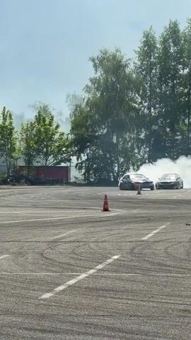 Two cars are drifting on a paved surface, creating large plumes of smoke. The car in the foreground has an orange, blue, and black design. The second car is lighter in color. There are several orange traffic cones on the ground. Trees and buildings are visible in the background under a bright sky.