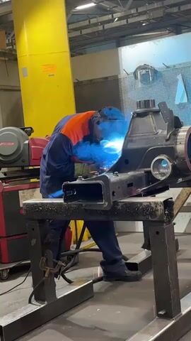 A person wearing a blue and orange jumpsuit and a welding helmet is welding a large metal object. The metal object appears to be part of a machine or vehicle component. The person is standing and bending over the object, which is supported by a metal stand. Sparks are flying from the welding torch. The environment appears to be a factory or workshop, with industrial equipment and yellow structures visible in the background.