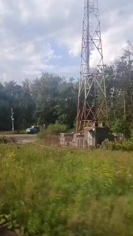 The video shows a view from a moving vehicle, likely a train, looking out at a rural or suburban landscape. In the foreground, tall green grass and yellow wildflowers are blurred due to motion. In the middle ground, a dirt road is visible with a blue truck and a person near it. To the right, a large metal tower, possibly a radio or cell tower, stands tall. Trees and bushes form a dense backdrop behind the road. The sky is partly cloudy. Electrical poles and wires are also visible along the roadside. The scene appears to be a roadside area with some industrial elements like the tower and electrical infrastructure interspersed with natural vegetation.