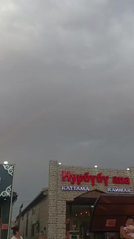 The video shows a view of a street with a building and mountains in the background. The building has red lettering that says "Нурбубу ана" with smaller lettering below that says "KATTAMA" and "КАЙМАК". There are several cars parked in front of the building and in a parking lot in the distance. The sky is cloudy with a rainbow visible in the upper left portion of the screen. There are people visible in the foreground, including a woman holding a baby.