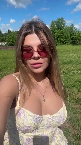 A young woman with long blonde hair and pink sunglasses is in an outdoor setting with green grass and trees in the background. She is wearing a light-colored, floral-patterned top. She smiles at the camera and then blows a kiss, holding her hand up to her mouth.