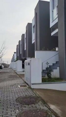 The video shows a row of modern townhouses with grey and white facades. A paved walkway made of interlocking bricks runs along the front of the houses. Each house has a set of stairs leading up to the entrance. Some houses have small landscaped areas with young plants. There are utility boxes along the walkway. The sky is overcast. A streetlight can be seen in the distance. The video appears to be taken from a low angle, moving slowly along the row of houses.