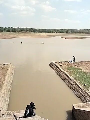 The video shows a body of water with several people visible. In the foreground, a person in a black hoodie sits on a stone structure. Further out in the water, two figures appear to be wading or fishing. On the right side of the frame, a person stands on a dry, rocky area near the edge of the water, with a blue bag beside them and a fishing rod in hand. The background shows a distant treeline and a hazy sky. The water is a murky brown color, and the land surrounding it appears dry and arid in places, suggesting a low water level.