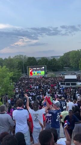 The video shows a large crowd of people gathered to watch a soccer game on a giant screen in an outdoor setting. The crowd appears excited, with many people cheering and waving flags. The screen is positioned in front of a backdrop of trees and a cityscape in the distance. The sky is overcast, suggesting it may be late afternoon or early evening. The crowd is diverse in age and ethnicity, and many are wearing jerseys or other sports-related attire.