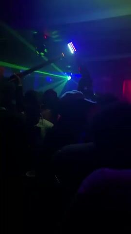The video shows a group of people in a club, likely dancing.  The lighting is dark, with colorful light effects.  Some individuals are holding light-up batons. The people are mostly silhouetted, making it hard to discern details. A person in a white top and another with a blue cap are visible amongst the crowd. The room appears crowded with people.