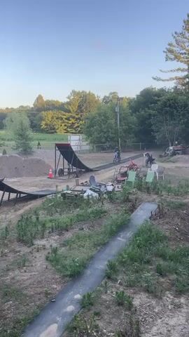 A motocross rider on a blue motorcycle performs a jump, launching off a ramp and soaring through the air. The rider is wearing a black helmet and riding gear. The jump occurs in an outdoor setting with trees and a clear blue sky in the background. The motorcycle lands on an inflatable airbag designed for extreme sports. The video shows the rider approaching the ramp, executing the jump, and landing safely.