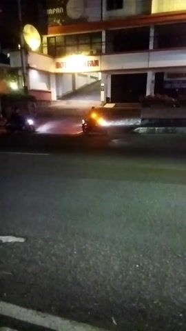 The video is a low-light, shaky recording of a street scene at night. In the foreground, a dark asphalt road is visible with some white markings. In the mid-ground, two motorcycles are present. The motorcycle on the left has its headlight on and appears to be stationary or moving slowly. The motorcycle on the right is in motion, with its rear lights illuminated and emitting a reddish-orange glow, possibly from exhaust or brake lights. There is also a bright yellow light emanating from the rear of this motorcycle. Behind the motorcycles, a building with a red and white facade is illuminated. The building has multiple windows and a prominent sign that is partially obscured but appears to read "MOTOFAN." The scene suggests an urban or semi-urban environment at night, possibly a street in front of a business. The lighting conditions create a dramatic effect with illuminated vehicles and building against the dark surroundings.