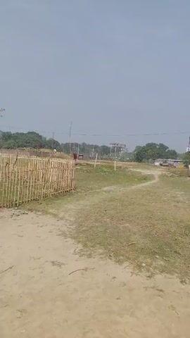 The video shows a dirt and grass field enclosed by a light-colored bamboo fence. A small, one-story building with a corrugated metal roof stands on the edge of the field, and a red flag flies atop the building. A single tree stands beside the building. The sky is clear and pale blue. In the distance, trees and buildings are visible. There are no people or animals in the immediate foreground. The camera pans slightly to the right. The overall impression is of a rural or agricultural setting.