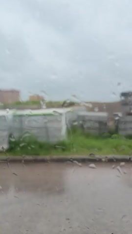 The video, filmed through a rain-streaked window, pans across an outdoor scene. In the foreground, the wet window obscures details, showing water droplets clinging to the glass. Beyond the window, a white car with a yellow triangular sticker on its rear is visible. The car is parked next to a patch of grass, with what appears to be a construction site in the background. The construction site features piles of dirt and debris, several pieces of heavy machinery, including an excavator, and what looks like a trailer covered with a grey tarp. The ground is wet, suggesting recent rain, and the sky is overcast. The camera movement is a slow, continuous pan to the right, revealing more of the construction site as it progresses. No people are clearly visible in the footage.