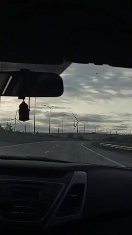 The video is filmed from the interior of a car, looking out the windshield. The car is traveling on a paved road that curves to the right. On either side of the road, there are fields with tall poles and what appear to be wind turbines. The sky is overcast with clouds. A black air freshener hangs from the rearview mirror. The dashboard of the car is visible in the foreground.