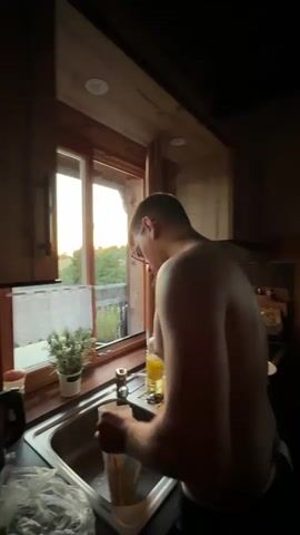 A person is standing at a kitchen sink, rinsing out a bag. The sink is stainless steel and there is a faucet. A window is to the left of the sink, showing greenery and a fence outside. The kitchen has wooden cabinets and a wooden countertop. The person is shirtless and appears to be male. The camera then pans to a stovetop with a pan on it. There are bottles and jars on a shelf above the stovetop. The camera then pans to a wooden table with plates, cutlery, and glasses on it. There is a television on the wall in the background. The room appears to be a rustic kitchen.