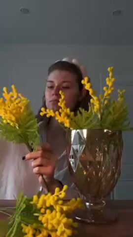 Here's a description of the video:

A woman with light hair and a headband is arranging faux yellow mimosa flowers in a gold-colored, textured glass vase. She is wearing a light-colored, striped pajama top. She holds a few stems in her left hand and is adjusting the foliage with her right. More sprigs of mimosa are on the wooden table in front of her. The background is a neutral-colored wall. A recessed light is visible in the ceiling.