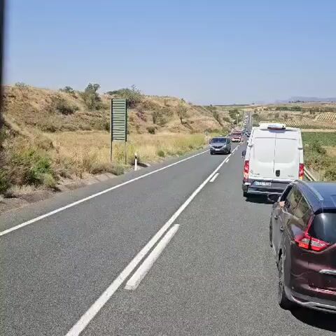 The video shows a traffic incident on a highway. Several vehicles are stopped on the right side of the road, including a fire truck, a white van, and several cars. A group of people, some wearing high-visibility vests, are gathered near the vehicles. One person is pointing into the distance. The highway is bordered by a dry, grassy hillside. The sky is clear and blue, indicating a sunny day. The overall scene suggests an emergency response or traffic management situation.