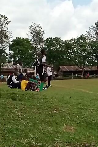 A medium shot depicts a soccer team on the sidelines of a field. Several players sit on a grassy bank in various team uniforms – blue and white, yellow, green. One player stands in a black and white uniform, pacing back and forth. A few adults stand near the players, dressed in white shirts. The background shows a green field, trees, and small buildings with tin roofs. The sky is partly cloudy.