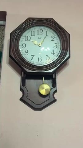 The video shows a wall-mounted pendulum clock with a wooden frame and a white face. The clock hands indicate the time is approximately 10:07.  The pendulum is brass-colored and hangs below the clock face. The wall is painted a light pink color. There is a window to the left of the clock.