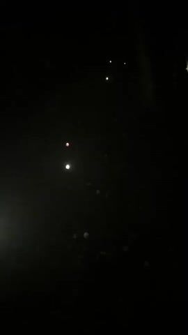 The video shows a dark outdoor scene at night. Multiple small, bright lights are visible, scattered across the ground and in the air. Some lights appear to be static, possibly from ground-level lamps or illuminated objects, while others seem to be moving or flickering, resembling fireflies or small, handheld lights. The environment appears to be a wooded area, with hints of trees and foliage in the background. The overall impression is of a magical or festive display of lights in a natural setting.