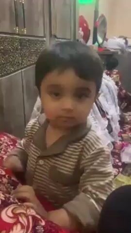 A young child sits on a bed covered with a red and black patterned blanket. The child is wearing a brown and beige striped pajama top. Behind the child is a wooden cabinet with decorative panels. The background is cluttered with various items, including a white ruffled garment and a mirror. The child looks directly at the camera with a serious expression. A person wearing black clothing is partially visible on the right side of the frame.