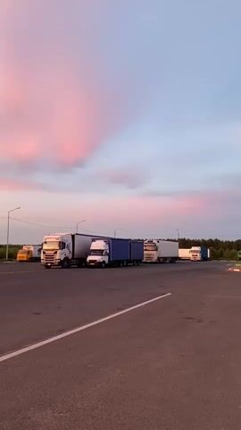 The video shows a parking lot with several semi-trailer trucks parked. The sky is filled with pink and blue clouds, suggesting it is either sunrise or sunset. The ground is paved asphalt with white lines marking parking spots. There are streetlights visible. The trucks are of various colors and designs, including white, blue, and yellow. Some trucks have trailers attached. There are no people visible in the video. The environment appears to be an outdoor rest stop or truck depot. The camera is static, facing a row of parked trucks. The lighting suggests it is either early morning or late evening.