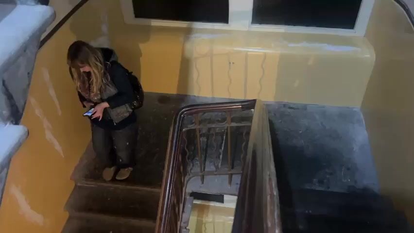 A person with long brown hair, wearing a dark vest and pants, walks down a stone staircase while looking at their phone. The walls are painted yellow and show signs of repair with white patches. A dark metal railing runs along the inside of the staircase. Above the landing is a small, dark window.