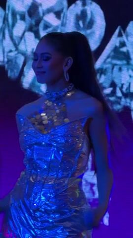 A woman with her hair in a ponytail is posing in a sparkly silver dress. She is wearing a jeweled necklace and earrings. The background is a dark purple with large, projected images of diamonds. The woman turns her head from side to side and then walks to the right, her dress shimmering under the lights.