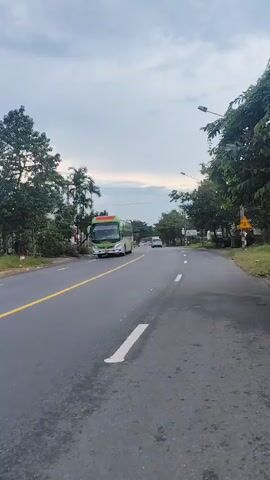 A video shows a bus driving on a road. The bus is green and white. There are trees on both sides of the road. The sky is cloudy. The road is asphalt and has yellow and white lines.
