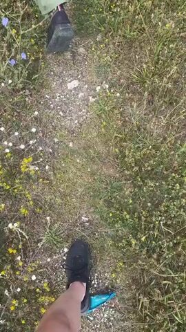 The video shows a person walking away from the camera on a dirt path through a field of grass and wildflowers. The person is wearing a green jumpsuit and black shoes. They are carrying a pair of white shoes and black socks in their left hand. Another person, whose legs and feet are visible in the foreground, is walking behind them on the same path. The second person is wearing black shoes and appears to be barefoot from the knee down. The sky is overcast, and trees are visible in the background.