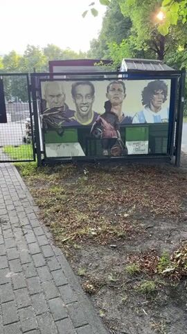 The video pans across a mural painted on a metal fence. The mural depicts four famous soccer players: Zinedine Zidane, Ronaldinho, Cristiano Ronaldo, and Diego Maradona. In the foreground, a silhouetted figure of a young person is painted, holding a soccer ball and a duffel bag. The figure is positioned in front of the mural of the soccer players, as if looking up at them. There is text written on the mural, partially obscured by the silhouetted figure, that appears to read "RESPECT FOR EVERYONE" and "RESPECT". The background shows green foliage and a building. The ground in front of the mural is covered with dry leaves and dirt, and a paved walkway is visible on the left side of the frame.