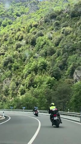 The video shows a winding road surrounded by a steep, green, tree-covered mountain. Several motorcyclists are riding along the road. One is wearing a bright yellow safety vest. There are blue and white road signs marking the curves of the road. The sky is blue with some white clouds. The camera appears to be positioned inside a vehicle following the motorcyclists.