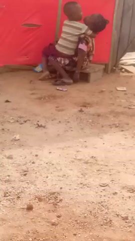 The video depicts a young child initially perched on an adult's back, near a structure with red covering. The child wears a striped shirt and maroon shorts, while the adult is dressed in a patterned top and skirt. They are sitting on a wooden block. As the video continues, the child dismounts and stands beside the seated adult. The setting appears to be a sandy area, with scattered debris like wrappers and a pair of blue Crocs nearby.