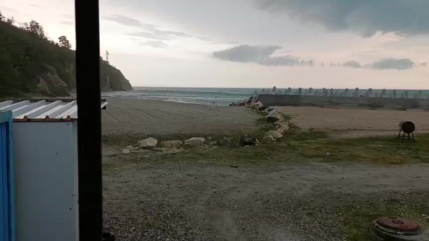 The video captures a panning view of a coastal scene in Byala, Bulgaria, during an overcast late afternoon or early evening.

The initial shot (0:00-0:02) looks out from an interior space, likely through a window or doorway, framing a sandy beach and the sea. On the left, a steep, forested cliff rises, while the sea is visible under a cloudy sky. In the mid-ground, a dirt path transitions to the sand, marked by scattered rocks. A concrete breakwater or pier extends into the sea on the right side of the frame. A dark, barrel-like object on a stand is situated on the dirt path.

As the camera pans left (0:02-0:05), it reveals more of the foreground structures. A prominent blue corrugated metal shed with a white corrugated roof is centered in the frame. Wires are visible across its roof. A similar white shed is partially visible behind and to the right of the blue one. The lush green cliff continues along the left background, while the beach and a glimpse of the sea remain visible on the right.

The scene depicts a functional coastal area, featuring natural elements like cliffs and the sea alongside utilitarian structures such as sheds and a breakwater. No people or visible text are present in the video.