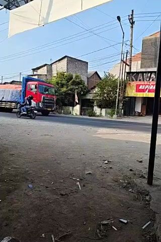 A red semi-truck with a blue tarp covering its cargo drives down a paved road. To the left of the truck are some buildings, one with a white awning. To the right of the truck is a street lamp and a building with a sign that says "KAWAN ASPIRASI". A black pickup truck drives past the semi-truck in the opposite direction. A person rides a motorcycle past the pickup truck.
