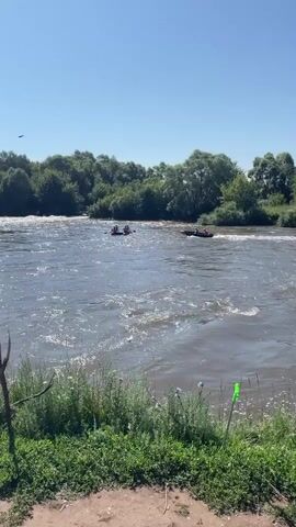 A wide shot captures a river with two rafts and a motorized boat. The rafts are positioned to the left of the frame, with multiple people visible in each. The motorized boat is to the right, moving at a moderate speed and creating a wake. The water appears choppy. The background consists of a dense line of green trees under a clear blue sky. The foreground shows grassy vegetation and a partially visible wooden stick. The scene suggests an outdoor recreational activity on a sunny day.