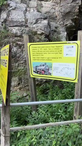 The video shows an outdoor environment with a large yellow sign on wooden posts in the foreground. The sign has text and an image of a steam train. Behind the sign, there is a rocky cliff face covered in greenery. To the right of the sign, a gray pole is visible with a red cylindrical object hanging from it. Further into the video, a rusty metal gate blocks the entrance to a tunnel. Several warning signs are attached to the gate. The tunnel appears dark and the surrounding area is overgrown with plants. The video pans slightly, giving a general view of the location.
