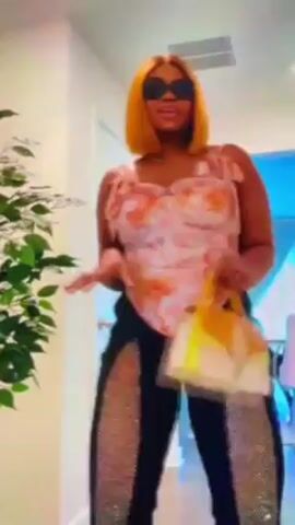 A woman with short blonde hair and dark sunglasses is standing and posing for the camera while holding a small yellow, white, and gold purse. She is wearing a light pink and orange floral corset and black pants with sparkling silver panels on the side. The scene is indoors with a plant to the left of the subject.