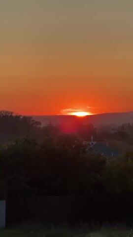 The video shows a sunset over a rural landscape. The sun is a bright orange orb low on the horizon, casting a warm glow across the sky. In the foreground, there are silhouetted trees and some houses with dark roofs. The overall scene is peaceful and serene, with a sense of the end of a day.