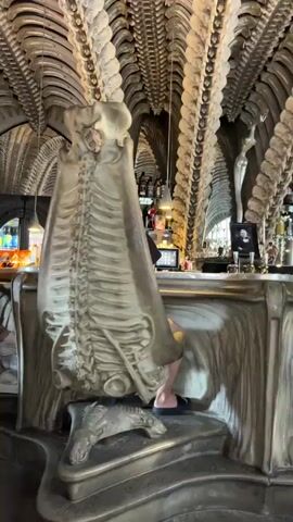 The video shows the interior of a bar with a biomechanical design, resembling the artwork of H.R. Giger. The architecture features ribbed, bone-like structures on the ceiling and walls, and the furniture, including chairs and the bar itself, are also designed in this style. In the foreground, a person is seated in a large, ornate chair that looks like a skeletal structure. The camera pans to the right, showing more of the bar area. Another person is seen sitting at the bar. Further to the right, another person is seated at a table, interacting with a laptop. The lighting is dim, with some spotlights highlighting the unique design elements. The overall impression is of a dark, surreal, and somewhat unsettling atmosphere.