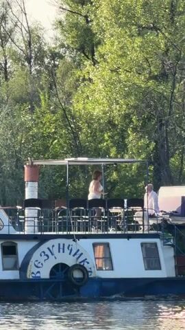 A boat with the inscription VEZYCHIK on its side is moving on a river. Two people are on the deck of the boat. A woman in a white swimsuit is dancing. A man in a white shirt and pants is standing to the right of the woman, watching her. The deck is covered with a canopy. There are chairs on the deck. The boat is surrounded by trees.
