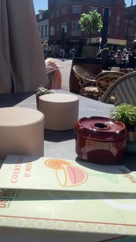 The video captures a scene at an outdoor café.  In the foreground is a table with three cylindrical beige objects, a red ceramic ashtray with a hole, a small potted plant, and a menu with a cocktail illustration.  The table is under the shade of a beige umbrella.  In the background, there's a view of a European-style town square.  A few people can be seen walking and cycling.  There are other café tables with patrons seated. A red brick building is visible across the square, and trees are planted in pots. The weather appears sunny.